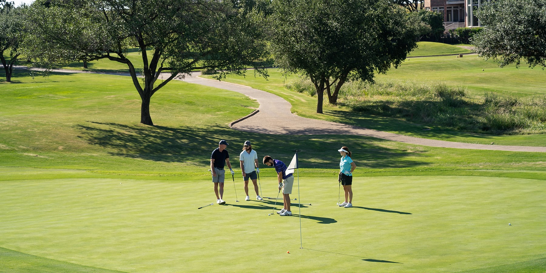 Four people stand on a sunny golf course green, with trees and a winding path in the background.