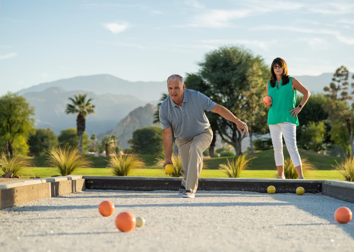 A man throws a bocce ball as a woman watches, with mountains and palm trees in the background.