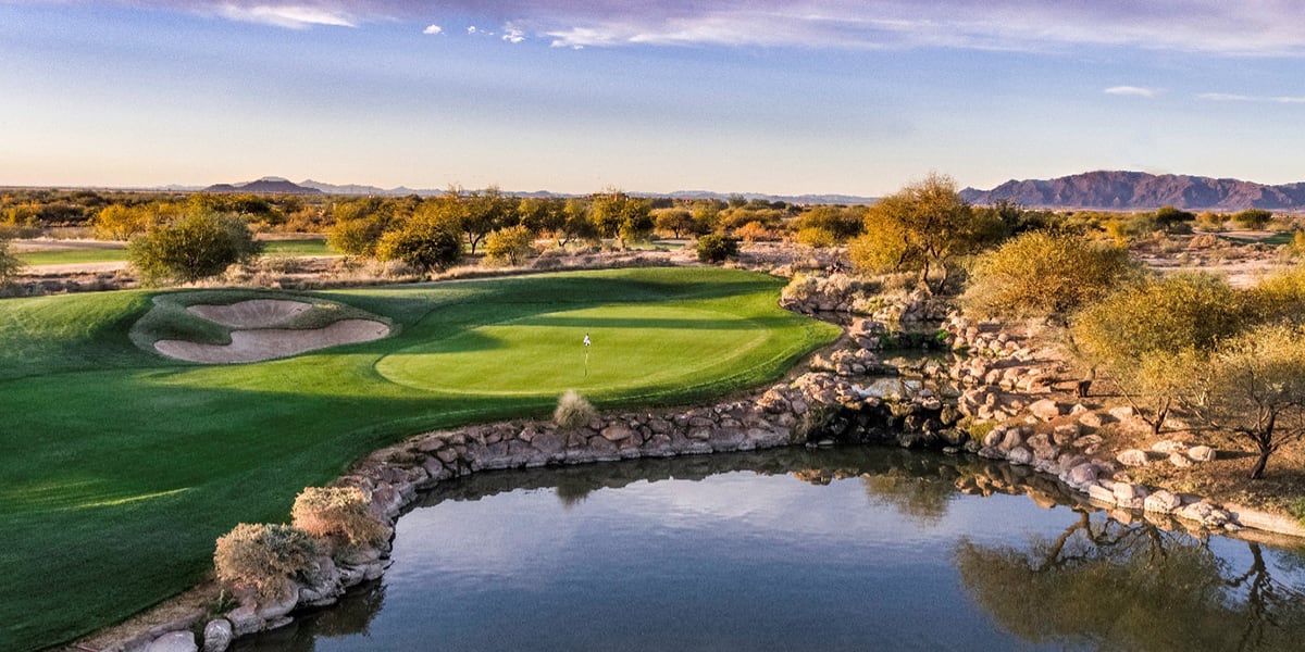 Golf course with green grass, sand bunker, pond, and desert landscape with mountains in the background.