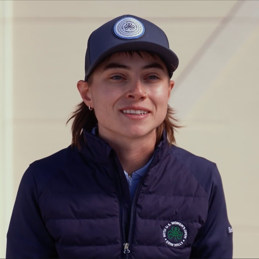 A smiling person in a navy jacket and cap with a golf club emblem, standing outdoors.