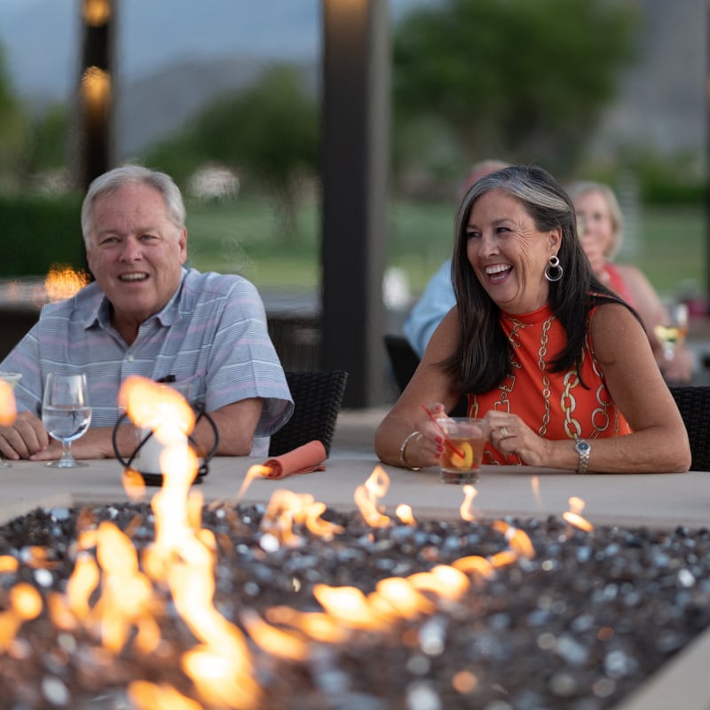Two people smile and relax by an outdoor fire pit, holding drinks at a patio table on a sunny day.