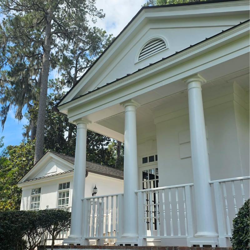 White house with tall columns and porch, surrounded by trees and greenery on a sunny day.